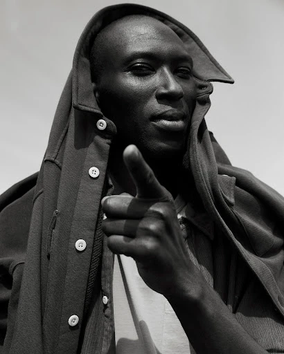 Monochromatic portrait of a Black adult male in a hooded jacket pointing directly at the camera, set against a plain, bright sky.