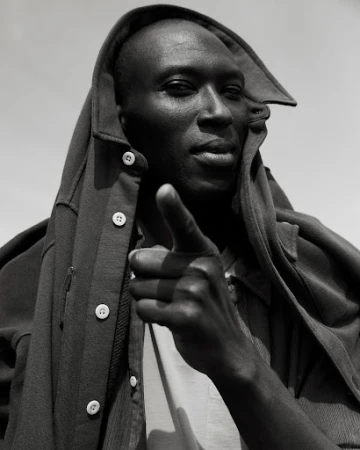 Monochromatic portrait of a Black adult male in a hooded jacket pointing directly at the camera, set against a plain, bright sky.