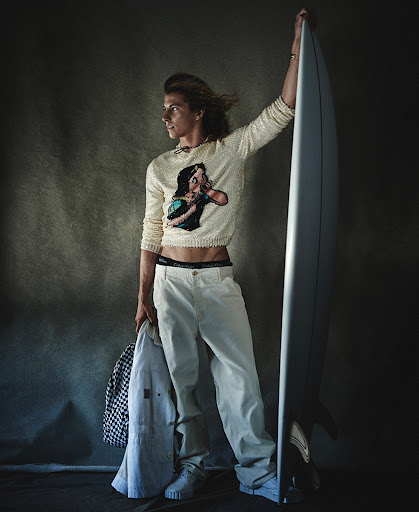 Young man with long hair holding a surfboard, wearing a graphic sweater, cream trousers, and white sneakers in a studio setting.