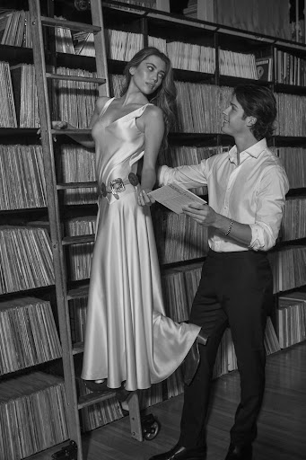 Black and white photo of a Caucasian woman in a dress standing on a ladder while a man in a shirt and pants holds papers nearby.