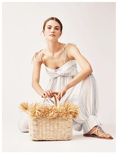 Woman in light blue and white striped jumpsuit poses with a straw bag in a bright, neutral studio. Fashion forward and fresh.