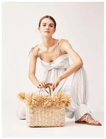 Woman in light blue and white striped jumpsuit poses with a straw bag in a bright, neutral studio. Fashion forward and fresh.