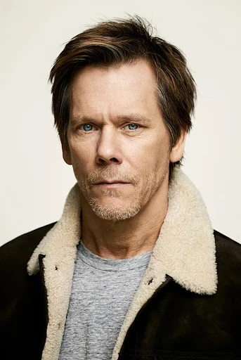 Close-up portrait of an adult male with brown hair and blue eyes wearing a jacket with a shearling collar and a gray shirt against a light background.