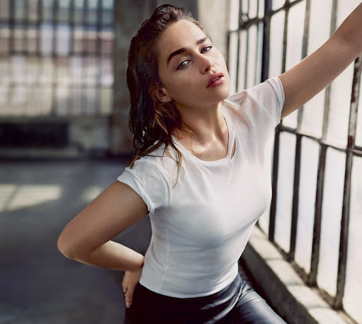Close-up of Emilia Clarke in a white tee and black pants near a window with blurred city background, emphasizing casual fashion and urban style.