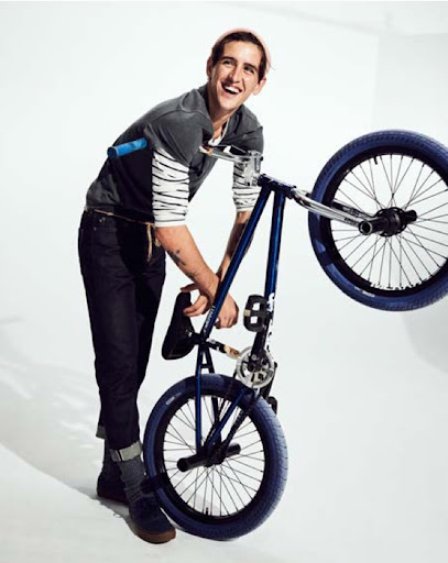 Man smiling next to a blue BMX bike indoors in a studio setting, captured in a medium shot with a bright, neutral backdrop.