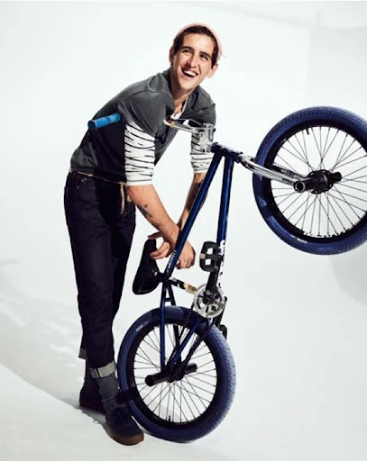 Man smiling next to a blue BMX bike indoors in a studio setting, captured in a medium shot with a bright, neutral backdrop.