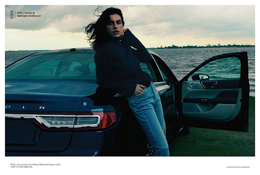 An adult woman leans against the trunk of a navy Lincoln Continental car, parked near a body of water under an overcast sky.