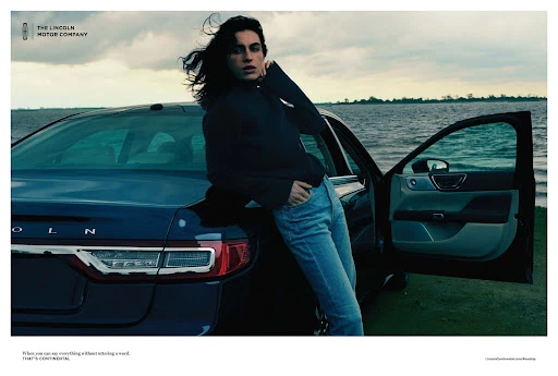 An adult woman leans against the trunk of a navy Lincoln Continental car, parked near a body of water under an overcast sky.