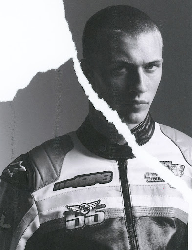 Monochrome portrait of a young adult male in a motorcycle jacket against a grey background, partially obscured by a torn paper effect.