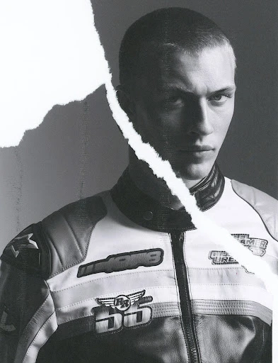 Monochrome portrait of a young adult male in a motorcycle jacket against a grey background, partially obscured by a torn paper effect.