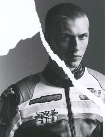 Monochrome portrait of a young adult male in a motorcycle jacket against a grey background, partially obscured by a torn paper effect.