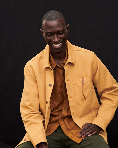 An African American man in a tan chore coat smiles warmly against a dark background, capturing a genuine and inviting moment.