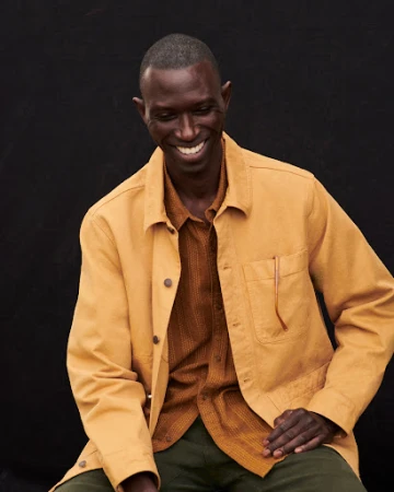 An African American man in a tan chore coat smiles warmly against a dark background, capturing a genuine and inviting moment.