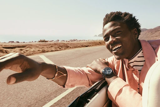 Smiling African American man enjoys a ride in a convertible car, displaying his wristwatch against an ocean and highway backdrop.