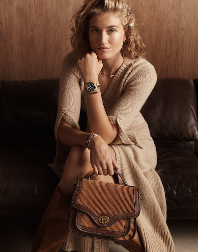 Portrait of a blonde adult woman sitting on a black couch wearing a beige dress, holding a brown purse, and a green wristwatch.