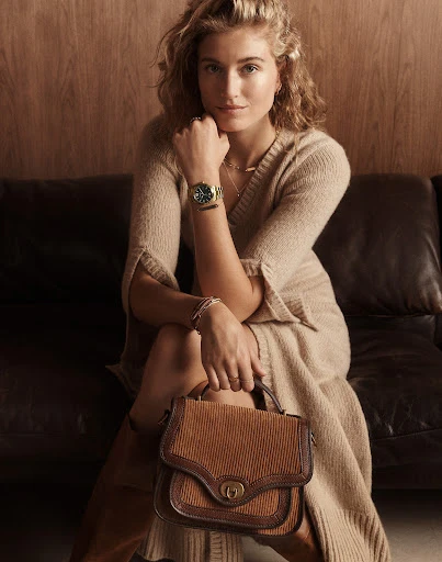 Portrait of a blonde adult woman sitting on a black couch wearing a beige dress, holding a brown purse, and a green wristwatch.