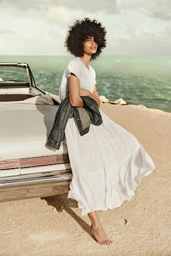 A woman with afro hair and white dress poses next to a vintage convertible car on a beach with ocean backdrop and soft, diffused lighting.