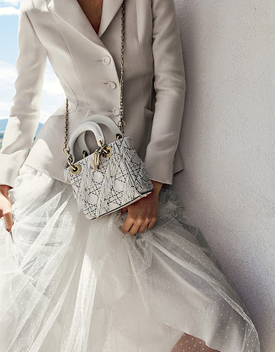 Woman in white blazer and tulle skirt poses with a Lady Dior handbag with chain strap on a white wall with blue sky.