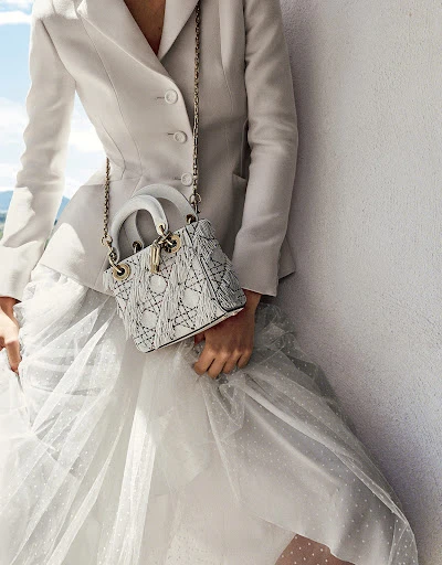 Woman in white blazer and tulle skirt poses with a Lady Dior handbag with chain strap on a white wall with blue sky.