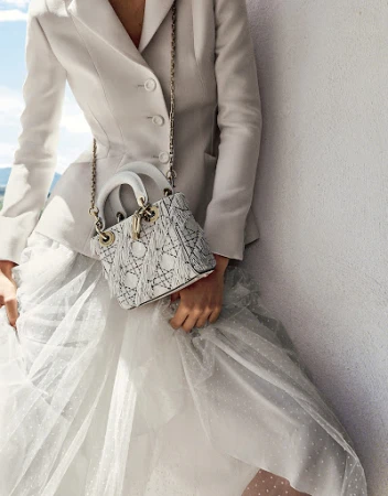 Woman in white blazer and tulle skirt poses with a Lady Dior handbag with chain strap on a white wall with blue sky.
