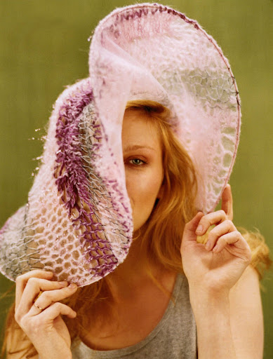 Portrait of a fair-skinned person wearing a pink and purple knitted hat covering one eye against a green backdrop, looking at camera.