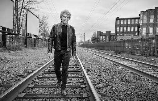 Black and white shot of an adult male walking on railroad tracks with industrial buildings in the background, under an overcast sky.