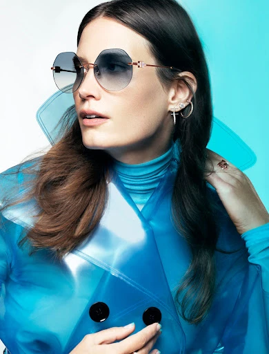 Portrait of a brunette woman wearing sunglasses and a translucent blue coat in a studio with gradient white to light blue background.