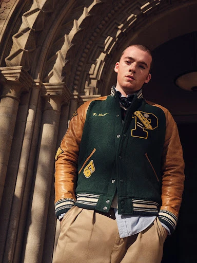 A young adult poses outdoors in collegiate jacket and khaki pants against an archway with stone pillars on a sunny day.