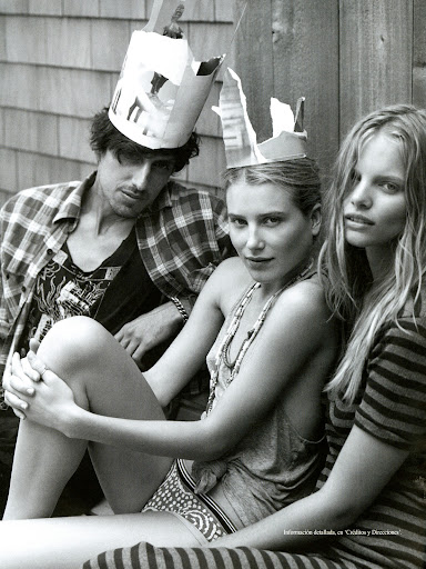 Black-and-white shot of a man and two women wearing paper crowns, posing in casual clothing in an outdoor setting with a wooden background.