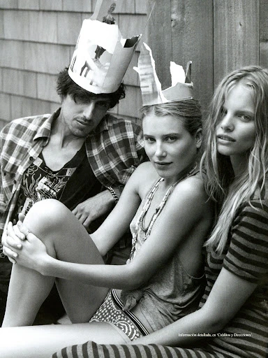 Black-and-white shot of a man and two women wearing paper crowns, posing in casual clothing in an outdoor setting with a wooden background.