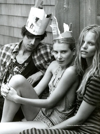 Black-and-white shot of a man and two women wearing paper crowns, posing in casual clothing in an outdoor setting with a wooden background.