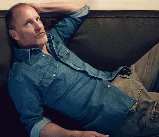 Adult male lying on a couch wearing a denim shirt and cargo pants looking at the camera in an indoor setting.  