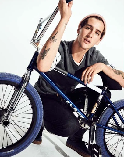 Man with tattoos poses next to a blue BMX bicycle in a brightly lit studio. He is wearing a beanie and dark clothing.