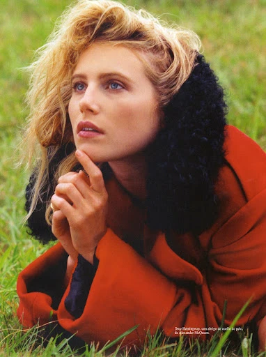 Portrait of a blond woman with blue eyes lying in grass wearing a red coat with a black fur collar looking up and to the side.
