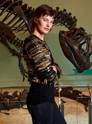 Adult female poses indoors wearing a decorative jacket. In the background is a dinosaur skeleton. Interior fashion shot in a museum.