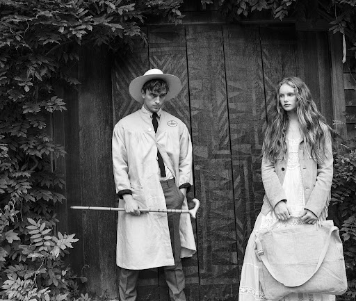 Monochrome fashion editorial featuring a man in a wide-brim hat holding a cane and a woman holding a large canvas bag.