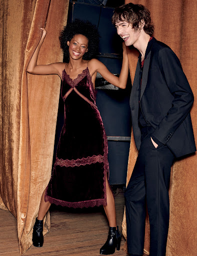 Fashion shot of an adult woman in a velvet dress and platform boots and an adult man in a suit standing near a curtain in a studio.