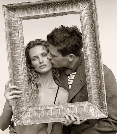 Monochrome portrait of a couple framed by an ornate picture frame; a man kisses a woman's cheek in a close-up shot.