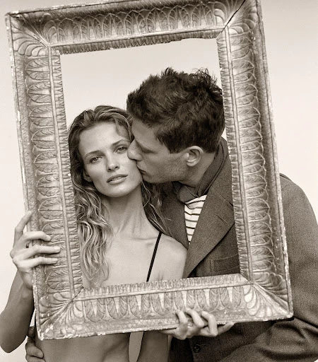 Monochrome portrait of a couple framed by an ornate picture frame; a man kisses a woman's cheek in a close-up shot.
