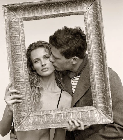 Monochrome portrait of a couple framed by an ornate picture frame; a man kisses a woman's cheek in a close-up shot.