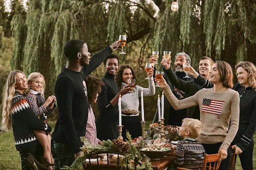 Family and friends gather around a rustic table outdoors, raising glasses in a toast, with a lush green backdrop and festive lighting.