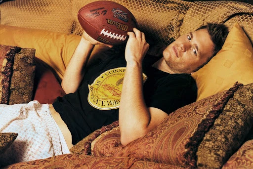 Man lying on a sofa holding a football wearing a black t-shirt and white patterned trousers styled by Deborah Watson