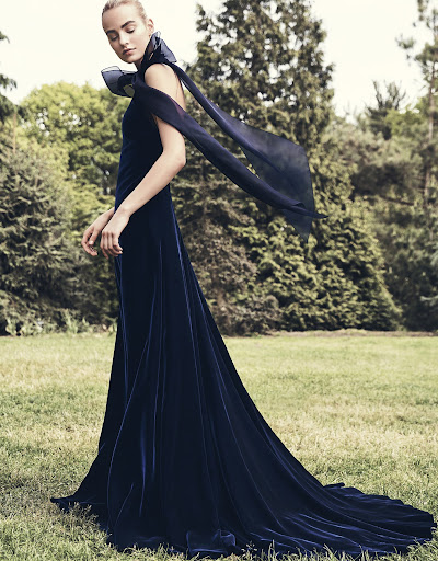 Elegant woman in a navy velvet gown with flowing scarf poses gracefully in a lush outdoor setting, blending fashion with nature.