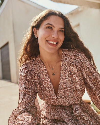 A woman with long brown hair poses indoors wearing a dark top and jeans against a neutral background, photographed with soft lighting.