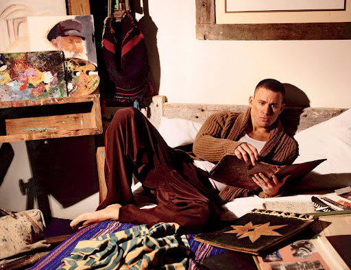 Man in cardigan lounging in bed, reading a book, surrounded by paintings and art supplies in a bedroom with natural lighting.
