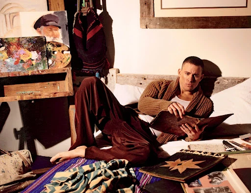 Man in cardigan lounging in bed, reading a book, surrounded by paintings and art supplies in a bedroom with natural lighting.