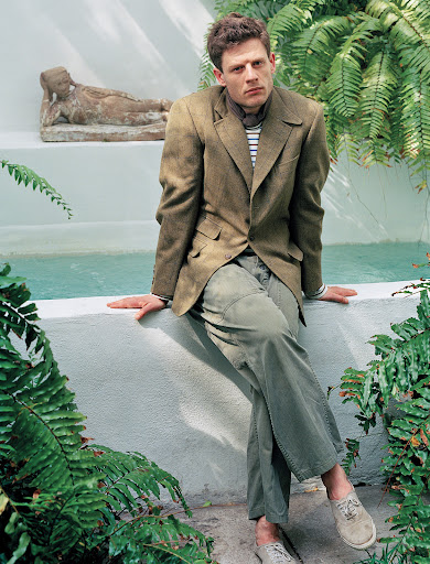 Male model in a brown jacket and green pants poses outdoors in front of a water feature and tropical foliage, looking directly at the camera.