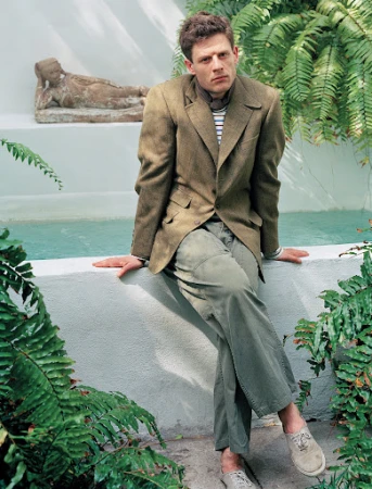 Male model in a brown jacket and green pants poses outdoors in front of a water feature and tropical foliage, looking directly at the camera.