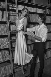 Black and white editorial image of a woman on a library ladder in a silk dress and a man holding a record standing beside her.