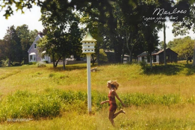 Woman running barefoot through a sunlit grassy field past a tall white birdhouse toward a country house.